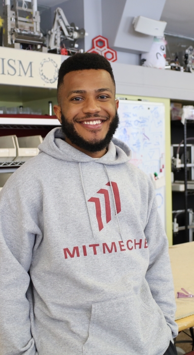 Headshot of Wilhem Hector leaning against a wood work table with a shelf filled with small organizers in the Pappalardo Lab at MIT